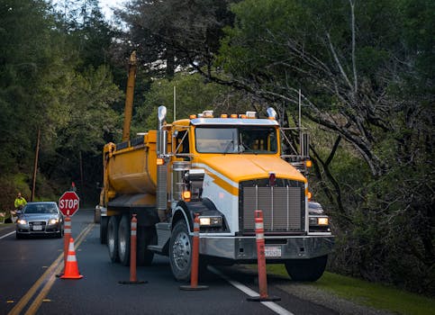 Yellow truck in road construction area surrounded by trees and traffic cones.