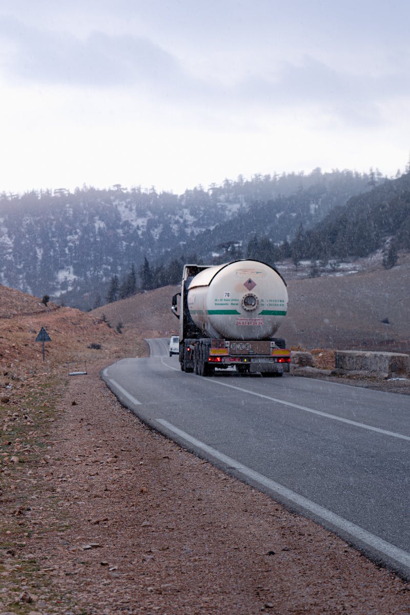 Gas tanker truck traveling on a mountainous road with snow-capped hills in the background.